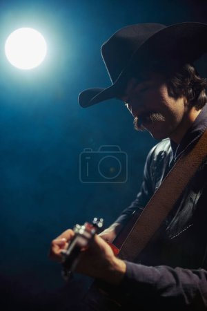 Photo for Portrait of man with moustaches in country style clothes playing guitar under spotlight isolated over dark blue background with smoke. Concept of music, creativity, inspiration, hobby, lifestyle - Royalty Free Image