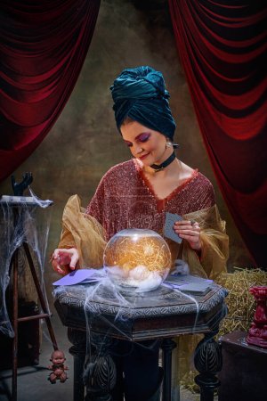 Photo for Portrait of magnificent fortune teller woman reading future on magical crystal ball and tarot cards over dark circus backstage background. Concept of vintage style, retro circus, art, fate - Royalty Free Image
