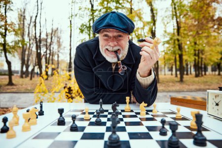 Photo for Portrait of delightful senior man playing chess in the park on a daytime in fall. Smoking pipe. Concept of leisure activity, friendship, sport, autumn season, game, entertainment, old generation - Royalty Free Image