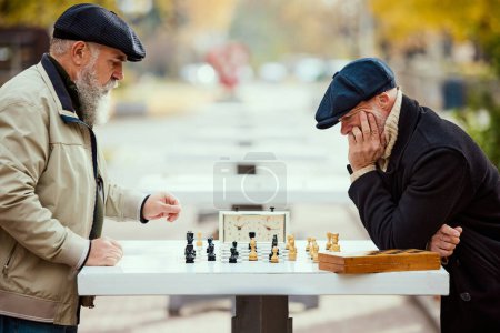 Téléchargez les photos : Portrait de deux hommes âgés jouant aux échecs dans le parc un jour d'automne. Une activité réfléchie. Concept d'activité de loisirs, amitié, sport, saison d'automne, jeu, divertissement, ancienne génération - en image libre de droit