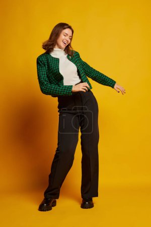 Portrait of young beautiful woman posing in stylish clothes, dancing isolated over yellow studio background. Concept of youth culture, emotions, facial expression, fashion. Copy space for ad