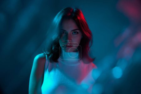 Portrait of young beautiful woman posing attentively looking at camera isolated over blue background in neon light. Concept of youth culture, emotions, facial expression, fashion. Copy space for ad