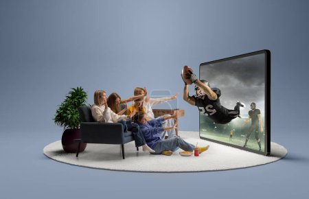Foto de Efecto de presencia. Jugador en vuelo. Sorprendidos jóvenes emocionados, niñas y niños sentados frente a un enorme modelo 3D de pantalla de TV en el interior de casa y viendo la emisión en línea del partido de fútbol americano - Imagen libre de derechos