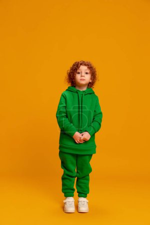 Photo for Portrait of cute little girl, child with curly red hair posing in green warm suit isolated over yellow background. Concept of childhood, emotions, lifestyle, fashion, happiness. Copy space for ad - Royalty Free Image