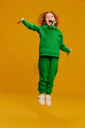 Téléchargez les photos : Portrait de mignonne petite fille, enfant aux cheveux roux bouclés sautant joyeusement et riant isolé sur fond jaune. Concept d'enfance, émotions, mode de vie, mode, bonheur. Espace de copie pour la publicité - en image libre de droit