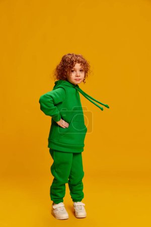 Photo for Portrait of cute little girl, child with curly red hair posing in green costume isolated over yellow background. Concept of childhood, emotions, lifestyle, fashion, happiness. Copy space for ad - Royalty Free Image