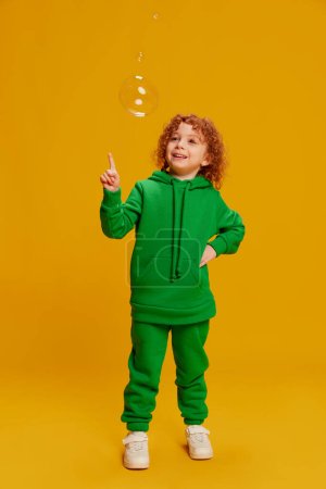 Photo for Portrait of cute little girl, child with curly red hair posing, playing with bubbles isolated over yellow background. Concept of childhood, emotions, lifestyle, fashion, happiness. Copy space for ad - Royalty Free Image