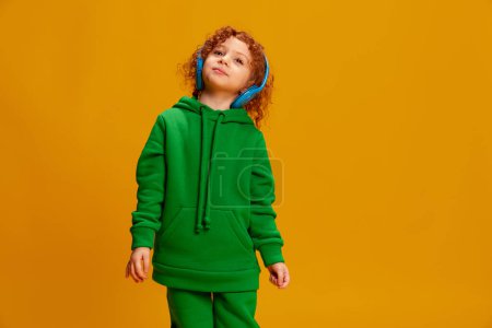 Photo for Portrait of cute little girl, child with curly red hair posing in headphones isolated over yellow background. Concept of childhood, emotions, lifestyle, fashion, happiness. Copy space for ad - Royalty Free Image
