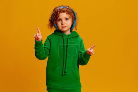 Photo for Portrait of cute little girl, child with curly red hair listening to music in headphones isolated on yellow background. Concept of childhood, emotions, lifestyle, fashion, happiness. Copy space for ad - Royalty Free Image
