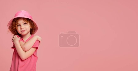 Téléchargez les photos : Portrait de mignonne petite fille, enfant aux cheveux roux bouclés posant en panama isolé sur fond rose. Le style. Concept d'enfance, émotions, mode de vie, mode, bonheur. Espace de copie pour la publicité - en image libre de droit