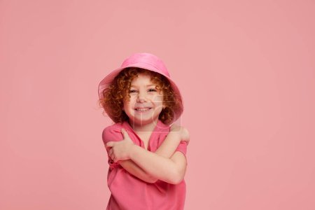 Photo for Portrait of cute little girl, child with curly red hair posing in panama and smiling isolated over pink background. Concept of childhood, emotions, lifestyle, fashion, happiness. Copy space for ad - Royalty Free Image