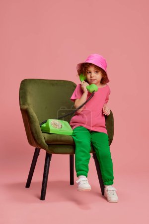 Photo for Portrait of cute little girl, child with curly red hair posing in panama, talking on phone isolated on pink background. Concept of childhood, emotions, lifestyle, fashion, happiness. Copy space for ad - Royalty Free Image