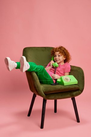 Photo for Portrait of cute little girl, child with curly red hair sitting on chair, talking on phone isolated on pink background. Concept of childhood, emotions, lifestyle, fashion, happiness. Copy space for ad - Royalty Free Image