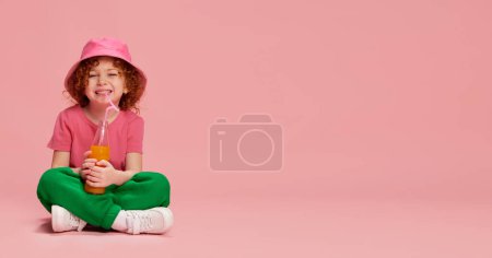 Téléchargez les photos : Portrait de mignonne petite fille, enfant aux cheveux roux bouclés assis au panama, buvant du jus isolé sur fond rose. Concept d'enfance, émotions, mode de vie, mode, bonheur. Espace de copie pour la publicité - en image libre de droit