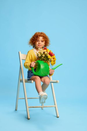 Téléchargez les photos : Portrait de mignonne petite fille, enfant aux cheveux roux bouclés posant sur chaise avec des fleurs isolées sur fond bleu. Concept d'enfance, émotions, mode de vie, mode, bonheur. Espace de copie pour la publicité - en image libre de droit