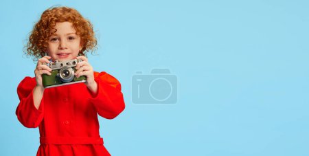 Téléchargez les photos : Petite fille mignonne, enfant aux cheveux roux bouclés posant, prenant des photos avec caméra vintage isolée sur fond bleu. Concept d'enfance, émotions, mode de vie, mode, bonheur. Espace de copie pour la publicité - en image libre de droit