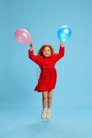 Photo for Portrait of cute little girl, child with curly red hair posing, jumping with balloons isolated over blue background. Concept of childhood, emotions, lifestyle, fashion, happiness. Copy space for ad - Royalty Free Image