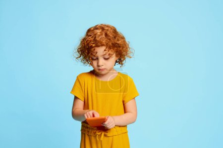 Photo for Portrait of cute little girl, child with curly red hair posing, playing on phone isolated over blue background. Concept of childhood, emotions, lifestyle, fashion, happiness. Copy space for ad - Royalty Free Image