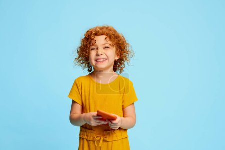 Photo for Portrait of cute little girl, child with curly red hair playing on phone, smiling isolated over blue background. Concept of childhood, emotions, lifestyle, fashion, happiness. Copy space for ad - Royalty Free Image