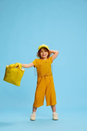 Photo for Portrait of cute little girl, child with curly red hair posing isolated over blue background. Stylish yellow outfit. Concept of childhood, emotions, lifestyle, fashion, happiness. Copy space for ad - Royalty Free Image