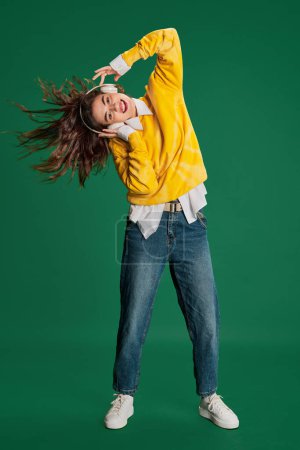 Foto de Retrato de niña alegre en suéter amarillo brillante, escuchando música en auriculares aislados sobre fondo verde. Concepto de juventud, belleza, moda, estilo de vida, emociones, expresión facial. Anuncio - Imagen libre de derechos