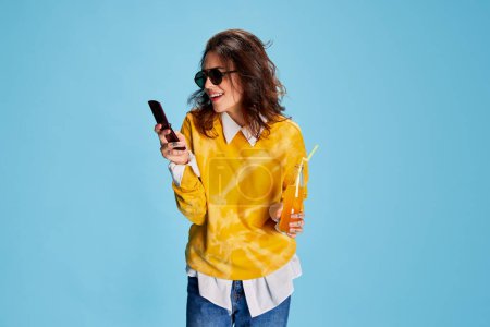 Photo for Portrait of young beautiful girl texting on phone and drinking juice isolated over blue background. Happy and delightful. Concept of youth, beauty, fashion, lifestyle, emotions, facial expression. Ad - Royalty Free Image