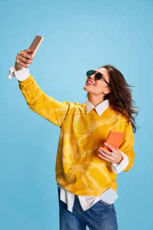 Foto de Joven hermosa chica sonriente en suéter amarillo brillante posando, tomando selfie con teléfono aislado sobre fondo azul. Concepto de juventud, belleza, moda, estilo de vida, emociones, expresión facial. Anuncio - Imagen libre de derechos