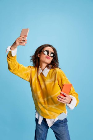 Foto de Joven chica hermosa posando en gafas de sol y suéter amarillo, tomando selfie con dos teléfonos aislados sobre fondo azul. Concepto de juventud, belleza, moda, estilo de vida, emociones, expresión facial. Anuncio - Imagen libre de derechos