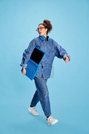 Photo for Portrait of young beautiful girl, student walking with laptop, posing isolated over blue background. Studying. Concept of youth, beauty, fashion, lifestyle, emotions, facial expression. Ad - Royalty Free Image