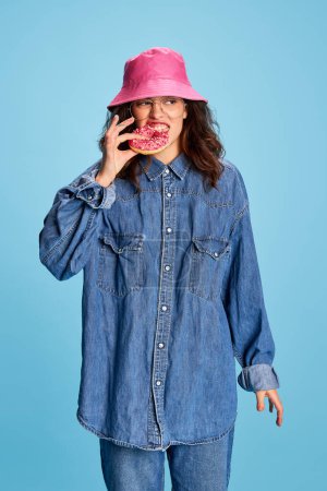 Photo for Portrait of young beautiful girl posing in pink panama, emotionally eating donut isolated over blue background. Concept of youth, beauty, fashion, lifestyle, emotions, facial expression. Ad - Royalty Free Image