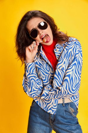 Photo for Portrait of young beautiful girl posing in stylish clothes and sunglasses, eating lollipop isolated on yellow background. Concept of youth, beauty, fashion, lifestyle, emotions, facial expression. Ad - Royalty Free Image