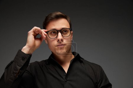 Photo for Serious young man, manager in black business classic style shirt putting eyewear on isolated over grey background. Fashion look of businessman. Concept of business, youth, modern lisfestyle. - Royalty Free Image