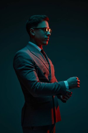 Photo for Fashionable serious young businessman in classic style suit adjusting his jacket sleeve isolated over dark background in neon light. Concept of business, fashion, style, modern lisfestyle. - Royalty Free Image