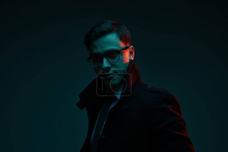 Photo for Studio shot of serious young 30s years old man in classic style clothes posing isolated over dark background in neon light. Fashion, style, emotions concept. Mental health, diversity, freedom - Royalty Free Image