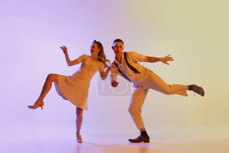 Téléchargez les photos : Belle fille et homme en costumes de style rétro dansant des danses incendiaires isolées sur fond dégradé de couleur lilas dans la lumière au néon. Danseurs en mouvement et en action. Concept d'art, culture des années 60, 70 - en image libre de droit