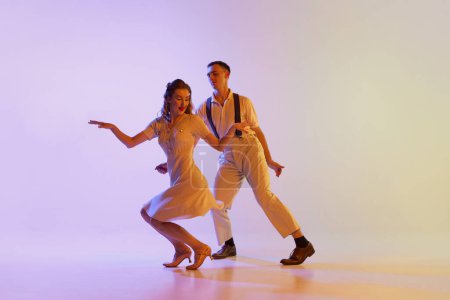 Musical show. Emotional couple of dancers in retro style outfits dancing social dances isolated on gradient lilac color background in neon light. Dancers in motion and action.