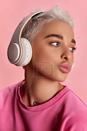 Photo for Young stylish woman with short hair and grey eyes listening to music in headphones, posing isolated over pink background. Concept of youth, beauty, fashion, lifestyle, emotions, facial expression. Ad - Royalty Free Image