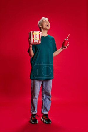 Photo for Portrait of young stylish woman with short hair posing with popcorn basket and laughing isolated over red background. Comedy. Concept of youth, beauty, fashion, lifestyle, emotions, facial expression - Royalty Free Image