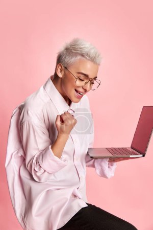 Photo for Portrait of young stylish woman with short hair, working on laptop in white shirt isolated over pink background. Successful deal. Concept of youth, beauty, fashion, lifestyle, emotions, business. Ad - Royalty Free Image