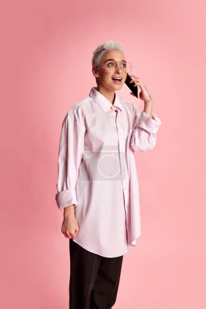 Photo for Portrait of young stylish woman with short hair posing in white shirt, talking on phone isolated over pink background. Concept of youth, beauty, fashion, lifestyle, emotions, facial expression. Ad - Royalty Free Image