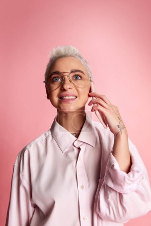 Photo for One young stylish woman with short hair posing in white shirt and glasses, talking on phone isolated over pink background. Concept of youth, beauty, fashion, lifestyle, emotions, facial expression. Ad - Royalty Free Image