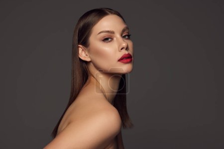 Photo for Female fashion model with naked shoulders and long hair looking at camera over dark grey background. Beautiful woman with trendy make-up, red lipstick and well-kept skin. Magazine style, beauty. - Royalty Free Image
