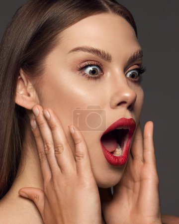 Foto de Retrato de cerca de una mujer joven y emotiva con maquillaje de moda, labios rojos posando con expresión impactada sobre un fondo gris oscuro. Concepto de belleza, moda, maquillaje, revista, emociones y publicidad. - Imagen libre de derechos