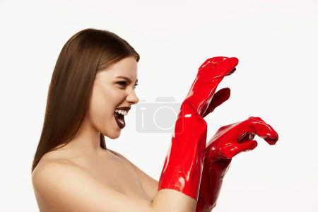 Photo for Portrait of beautiful young woman with straight brown hair and dark makeup posing in red rubber gloves on white background. Roaring. Party. Concept of beauty, fashion, emotions, magazing style and ad - Royalty Free Image