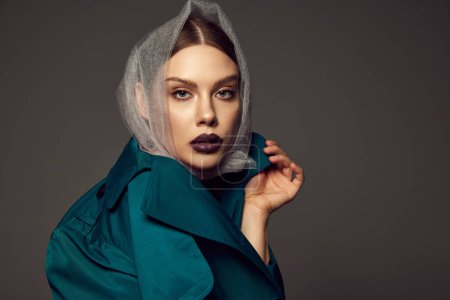 Photo for Portrait of young stylish woman with trendy dark lips make-up posing in fashionable coat and transparent headscarf over dark grey background. Concept of style, beauty, high fashion, magazine style, ad - Royalty Free Image