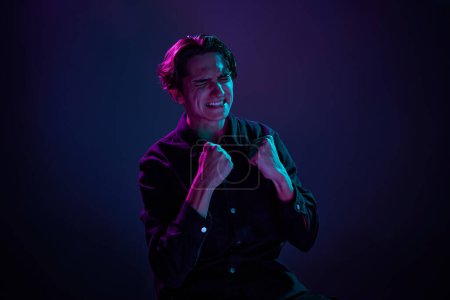 Photo for Portrait of young man in casual black shirt isolated over gradient dark purple background in neon light. Happy and excited. Concept of human emotions, facial expression, sales, ad, fashion and beauty - Royalty Free Image