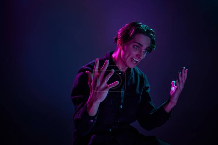 Photo for Portrait of young man in casual black shirt emotively posing isolated over gradient dark purple background in neon light. Concept of human emotions, facial expression, sales, ad, fashion and beauty - Royalty Free Image
