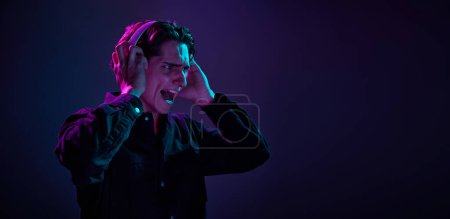 Foto de Retrato de hombre joven en camisa negra casual aislado sobre el gradiente fondo púrpura oscuro en luz de neón. Música en auriculares. Concepto de emociones humanas, expresión facial, ventas, publicidad, moda y belleza - Imagen libre de derechos