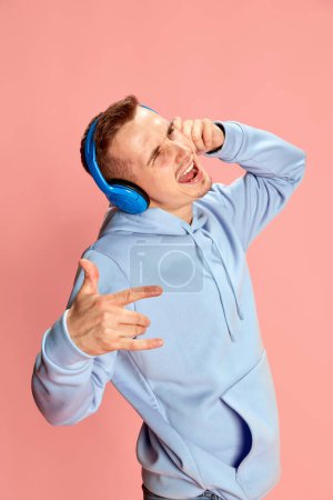 Foto de Retrato del joven hombre emotivo con capucha escuchando música en auriculares y cantando aislado sobre fondo rosa. Concepto de juventud, estilo de vida, música, moda casual, emociones, expresión facial. Anuncio - Imagen libre de derechos