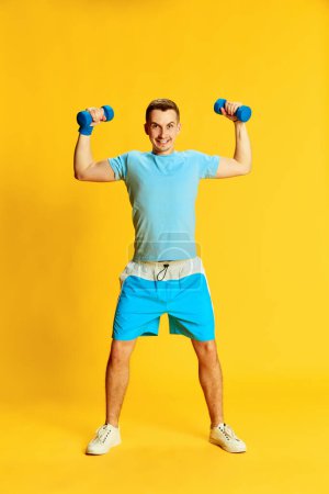 Foto de Retrato de un joven en uniforme de entrenamiento azul, haciendo ejercicios de manos con pesas, posando aislado sobre fondo amarillo. Concepto de deporte, estilo de vida fitness, cuidado corporal, salud, juventud, acción. Anuncio - Imagen libre de derechos